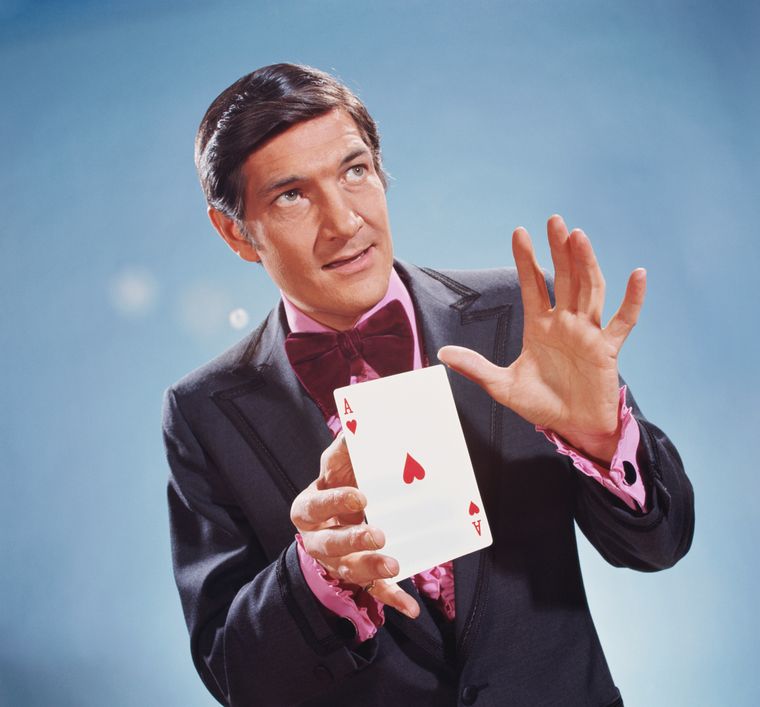 A magician in a vintage suit holds a large Ace of Hearts card, performing a dramatic trick against a blue background.